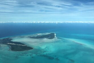 High-altitude aerial photograph of Bimini, Bahamas. Two main tropical islands are separated by a wide expanse of pale, translucent turquoise water and sandbars. The land is covered in dark green vegetation with visible developments and a small airstrip. A line of white fluffy clouds sits low on the horizon under a pale blue, wispy sky, meeting the deep blue edge of the deep ocean.