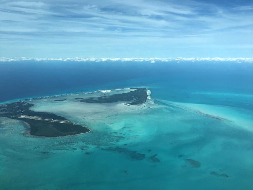 High-altitude aerial photograph of Bimini, Bahamas. Two main tropical islands are separated by a wide expanse of pale, translucent turquoise water and sandbars. The land is covered in dark green vegetation with visible developments and a small airstrip. A line of white fluffy clouds sits low on the horizon under a pale blue, wispy sky, meeting the deep blue edge of the deep ocean.