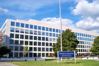 Wide view of a large, modern office building with a white stone façade and rows of square, reflective windows arranged in a grid pattern. In the foreground, a blue sign reads “Department of Transportation, Federal Aviation Administration, Wilbur Wright Building,” set on a neatly maintained grass lawn with small trees and pathways. A tall flagpole stands near the centre of the frame, while a pedestrian walkway runs along the left side. The scene is brightly lit under a blue sky with scattered white clouds, reflecting in the building’s glass windows.