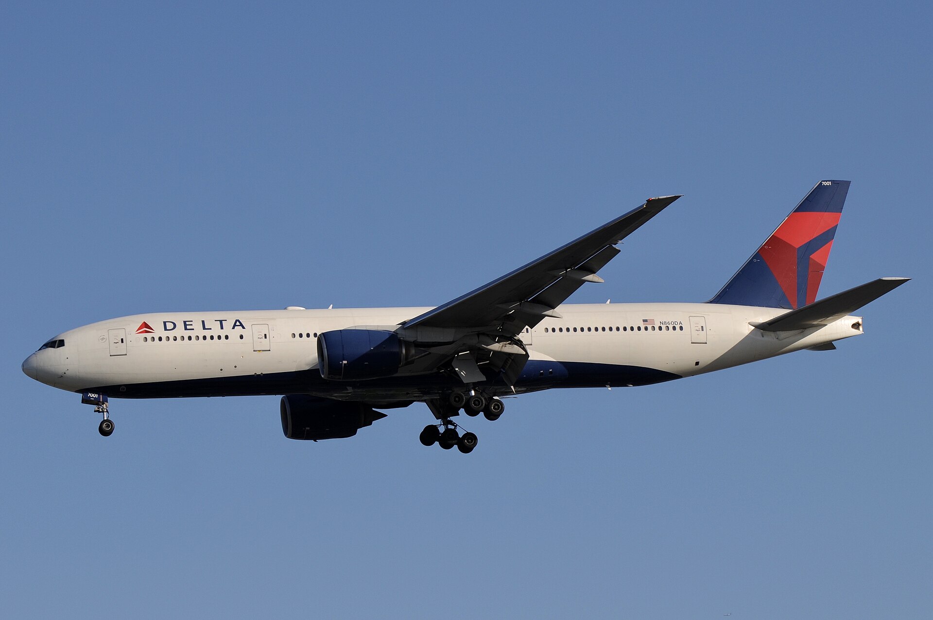 A low-angle shot of a Delta Air Lines Boeing 777 wide-body aircraft in flight. The plane has a white fuselage with a dark blue underbelly and the Delta logo near the nose. 