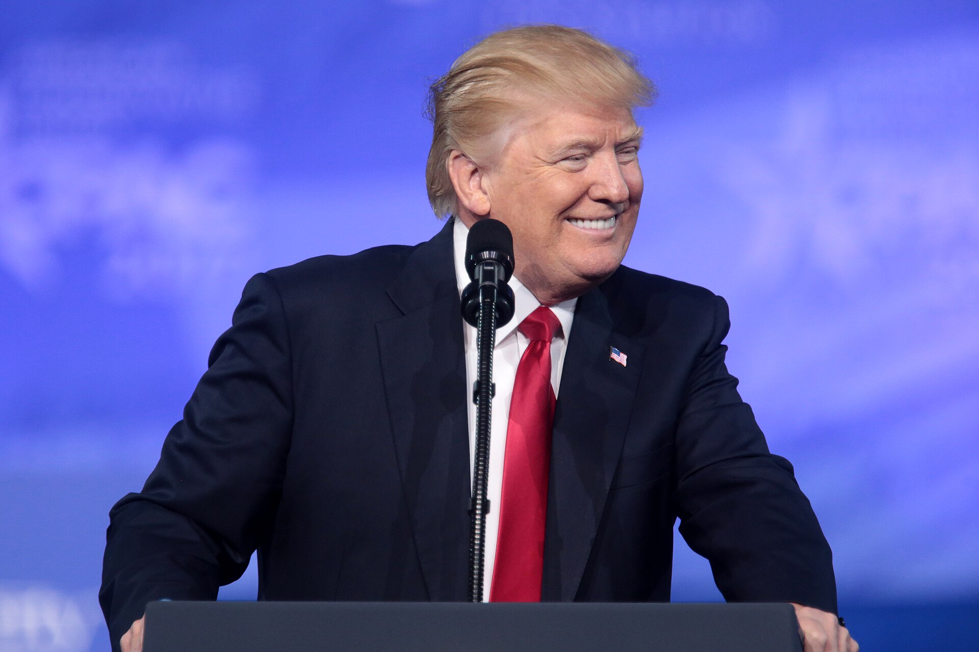 A medium shot of Donald Trump standing at a dark podium, smiling and looking toward his left. He is wearing a navy blue suit jacket, a white collared shirt, and a vibrant red necktie. A microphone is positioned in front of him, and a blurred blue background with white text and graphics is visible behind him.