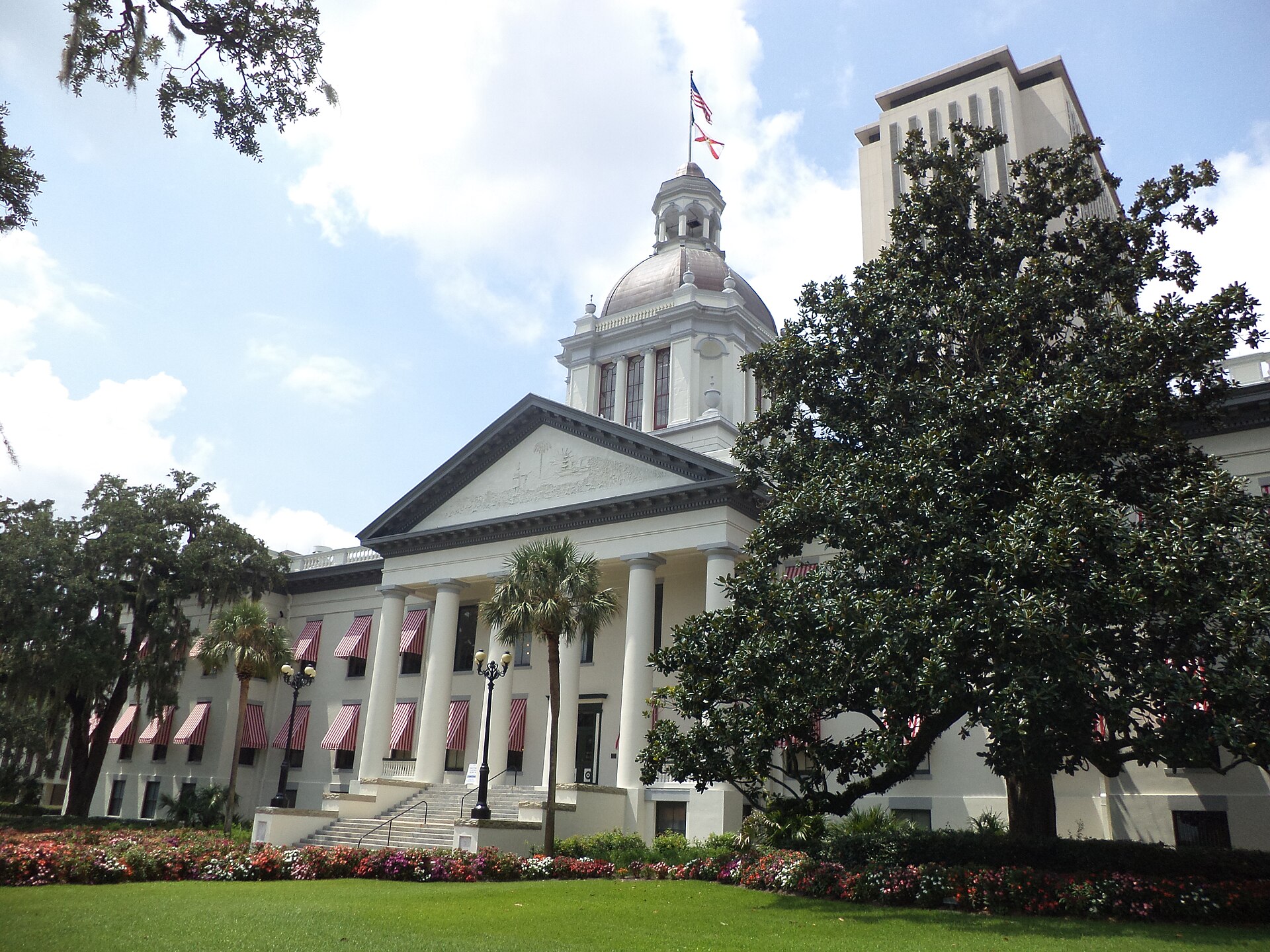 A wide-angle, low-reflection shot of the Florida State Capitol complex in Tallahassee. In the foreground is the historic Old Capitol, a white Neoclassical building with a prominent silver dome, red-and-white striped awnings over the windows, and a grand portico with tall columns. To the right and behind it rises the modern 22-story Capitol executive skyscraper. The scene is framed by lush green lawns, colorful flower beds, and large oak and palm trees under a partly cloudy sky.