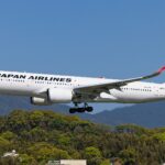 A side view of a Japan Airlines Airbus A350-1000 in mid-air with its landing gear deployed. The aircraft features a white fuselage with "JAPAN AIRLINES" written in black and the iconic red "Tsurumaru" crane logo on the tail, flying over a lush green hillside.
