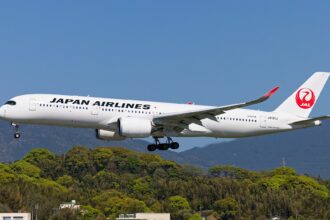 A side view of a Japan Airlines Airbus A350-1000 in mid-air with its landing gear deployed. The aircraft features a white fuselage with "JAPAN AIRLINES" written in black and the iconic red "Tsurumaru" crane logo on the tail, flying over a lush green hillside.