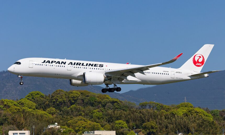 A side view of a Japan Airlines Airbus A350-1000 in mid-air with its landing gear deployed. The aircraft features a white fuselage with "JAPAN AIRLINES" written in black and the iconic red "Tsurumaru" crane logo on the tail, flying over a lush green hillside.