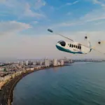 Mehair aircraft flying over the sea with a city in the left background.