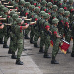 A high-angle, medium shot shows a large group of Mexican soldiers in green digital camouflage uniforms and helmets. Several soldiers in the foreground are playing gold-colored bugles, while others hold drums decorated with red and gold fringe. Many of the soldiers wear distinctive red braided cords on their upper sleeves, and all are positioned in an organized formation on a grey stone surface.