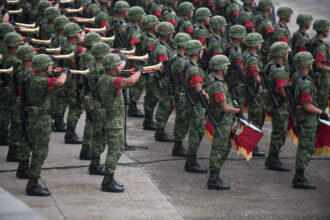 A high-angle, medium shot shows a large group of Mexican soldiers in green digital camouflage uniforms and helmets. Several soldiers in the foreground are playing gold-colored bugles, while others hold drums decorated with red and gold fringe. Many of the soldiers wear distinctive red braided cords on their upper sleeves, and all are positioned in an organized formation on a grey stone surface.