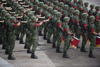 A high-angle, medium shot shows a large group of Mexican soldiers in green digital camouflage uniforms and helmets. Several soldiers in the foreground are playing gold-colored bugles, while others hold drums decorated with red and gold fringe. Many of the soldiers wear distinctive red braided cords on their upper sleeves, and all are positioned in an organized formation on a grey stone surface.