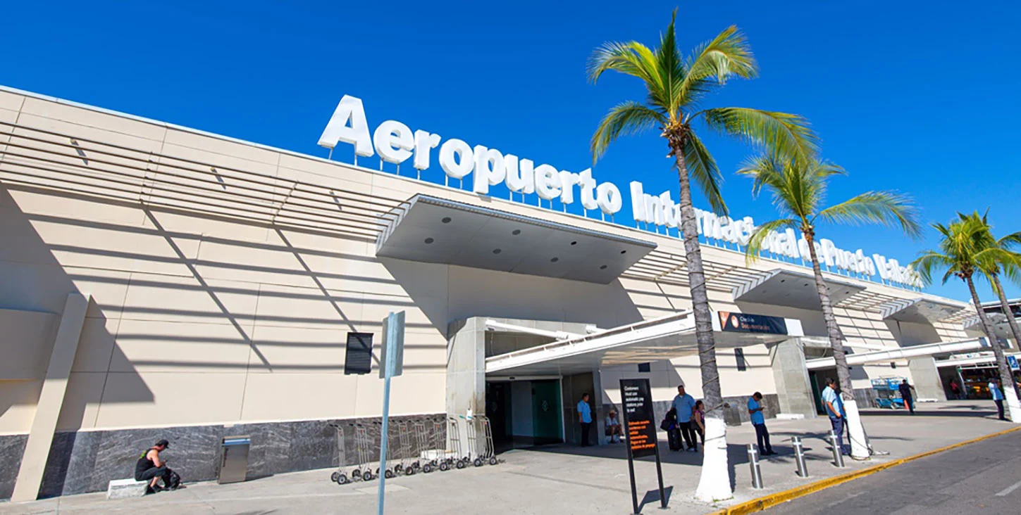 A wide, low-angle shot of the modern exterior of the Puerto Vallarta International Airport (Aeropuerto Internacional de Puerto Vallarta) in Mexico. Large white 3D letters spell out the airport's name across the top of a tan building under a bright blue sky. Tall palm trees line the sidewalk in front of the entrance, where travelers and airport staff are gathered near baggage carts and terminal doors.