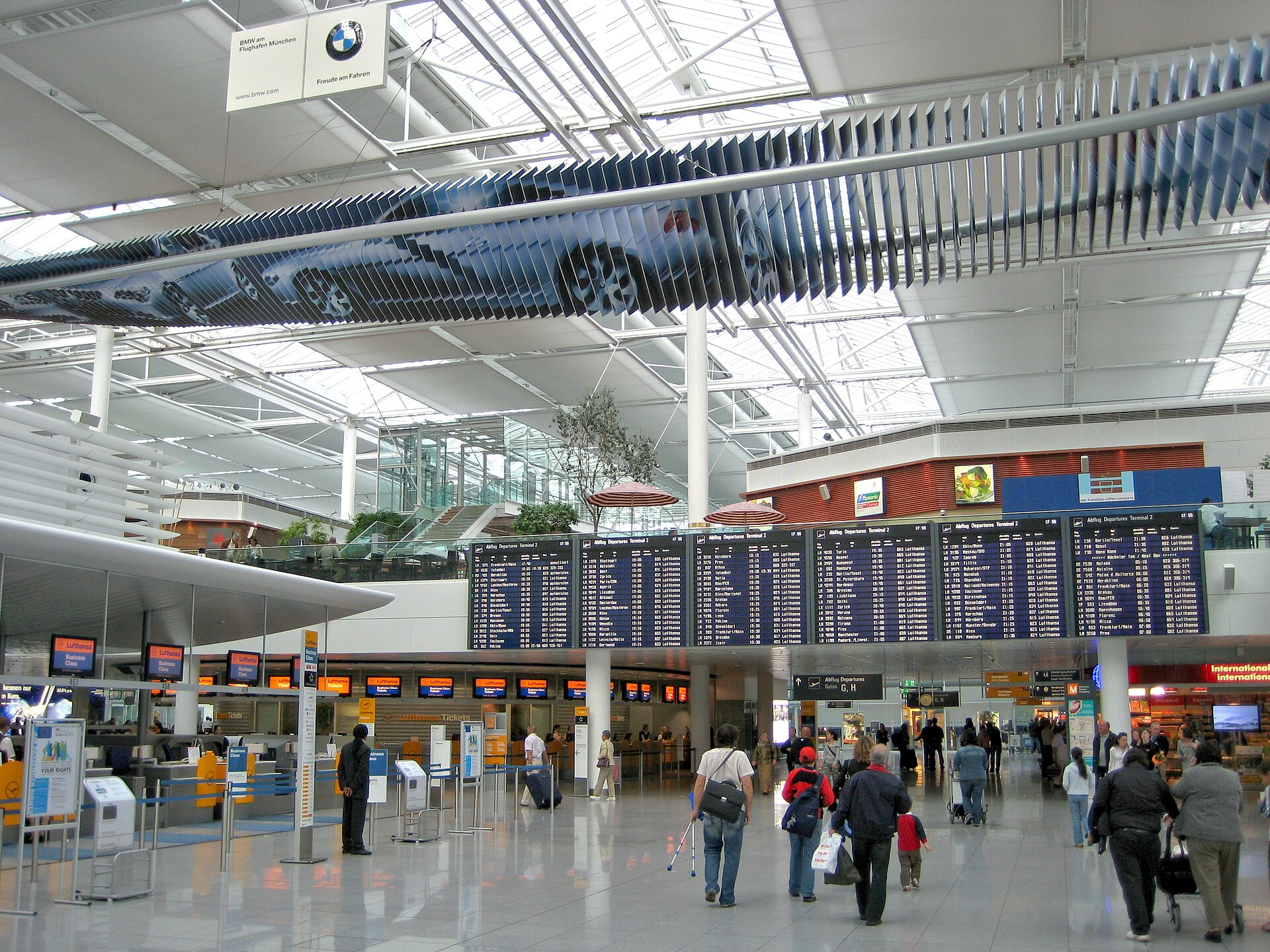 A wide-angle interior photograph of a modern airport terminal. In the center, several large, blue electronic flight departure boards hang above the concourse. Above them, a long, kinetic art installation consisting of numerous vertical slats creates the image of a car. To the left, several Lufthansa business class check-in counters are visible. People with luggage walk across the polished, reflective grey floor. The space is bright, with a high ceiling supported by white pillars and a large skylight structure that lets in natural light.