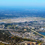 An aerial, high-angle photograph showing Palm Beach International Airport (PBI) and the surrounding urban landscape of West Palm Beach, Florida, under a clear blue sky. The airport's runways and taxiways dominate the center of the frame, bordered by dense residential neighborhoods, commercial buildings, and major highway interchanges. A large blue lake is visible in the lower right, and the Atlantic Ocean horizon stretches across the background.