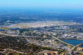 An aerial, high-angle photograph showing Palm Beach International Airport (PBI) and the surrounding urban landscape of West Palm Beach, Florida, under a clear blue sky. The airport's runways and taxiways dominate the center of the frame, bordered by dense residential neighborhoods, commercial buildings, and major highway interchanges. A large blue lake is visible in the lower right, and the Atlantic Ocean horizon stretches across the background.