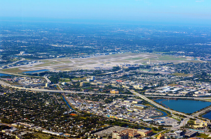 An aerial, high-angle photograph showing Palm Beach International Airport (PBI) and the surrounding urban landscape of West Palm Beach, Florida, under a clear blue sky. The airport's runways and taxiways dominate the center of the frame, bordered by dense residential neighborhoods, commercial buildings, and major highway interchanges. A large blue lake is visible in the lower right, and the Atlantic Ocean horizon stretches across the background.