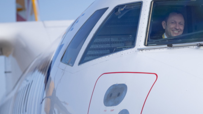 A smiling man in the pilots seat captured through a window on a Aurigny plane
