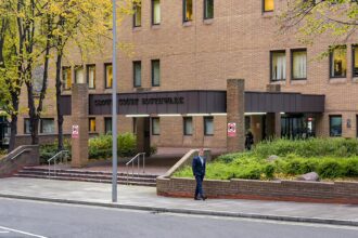 A wide-angle shot of the brick exterior of the Southwark Crown Court building. The entrance features a large overhang with the words "CROWN COURT SOUTHWARK" in dark lettering. A man in a blue suit walks along the sidewalk in the foreground, while autumn trees and green shrubbery surround the brick structure.