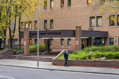 A wide-angle shot of the brick exterior of the Southwark Crown Court building. The entrance features a large overhang with the words "CROWN COURT SOUTHWARK" in dark lettering. A man in a blue suit walks along the sidewalk in the foreground, while autumn trees and green shrubbery surround the brick structure.