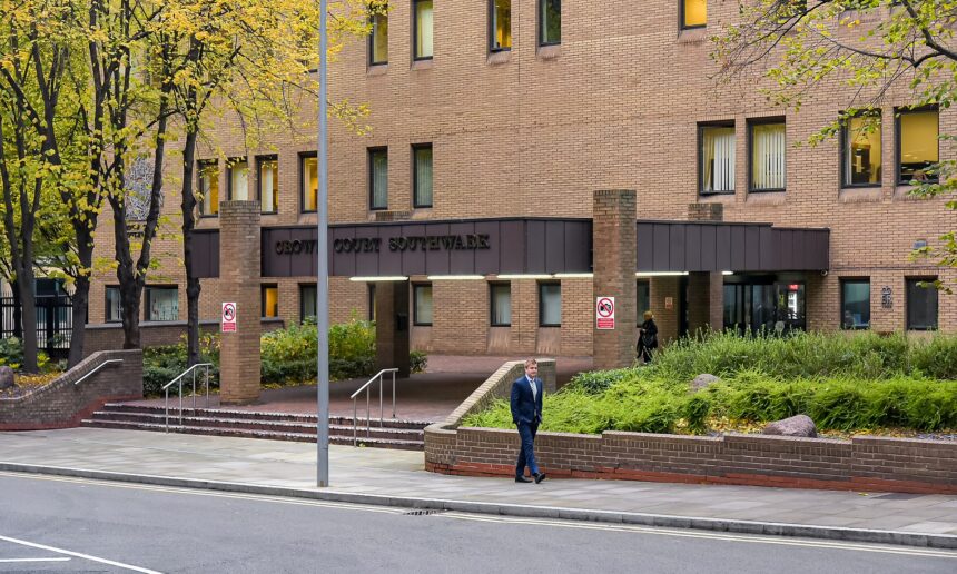 A wide-angle shot of the brick exterior of the Southwark Crown Court building. The entrance features a large overhang with the words "CROWN COURT SOUTHWARK" in dark lettering. A man in a blue suit walks along the sidewalk in the foreground, while autumn trees and green shrubbery surround the brick structure.