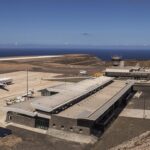 A high-angle, wide shot of St Helena Airport (HLE), showcasing its unique location on a leveled clifftop surrounded by the deep blue South Atlantic Ocean. The modern terminal building sits near the runway, which was constructed across the "Dry Gut" fill. The rugged, rocky terrain of the island’s coastline is visible under a clear sky.