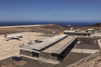 A high-angle, wide shot of St Helena Airport (HLE), showcasing its unique location on a leveled clifftop surrounded by the deep blue South Atlantic Ocean. The modern terminal building sits near the runway, which was constructed across the "Dry Gut" fill. The rugged, rocky terrain of the island’s coastline is visible under a clear sky.