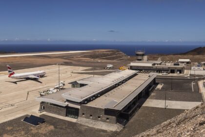 A high-angle, wide shot of St Helena Airport (HLE), showcasing its unique location on a leveled clifftop surrounded by the deep blue South Atlantic Ocean. The modern terminal building sits near the runway, which was constructed across the "Dry Gut" fill. The rugged, rocky terrain of the island’s coastline is visible under a clear sky.