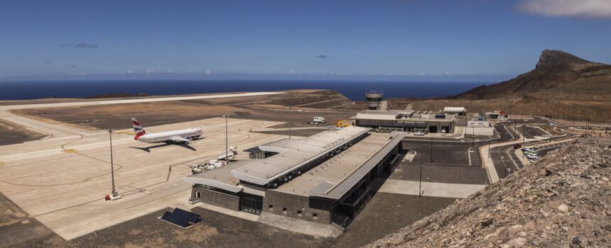 A high-angle, wide shot of St Helena Airport (HLE), showcasing its unique location on a leveled clifftop surrounded by the deep blue South Atlantic Ocean. The modern terminal building sits near the runway, which was constructed across the "Dry Gut" fill. The rugged, rocky terrain of the island’s coastline is visible under a clear sky.
