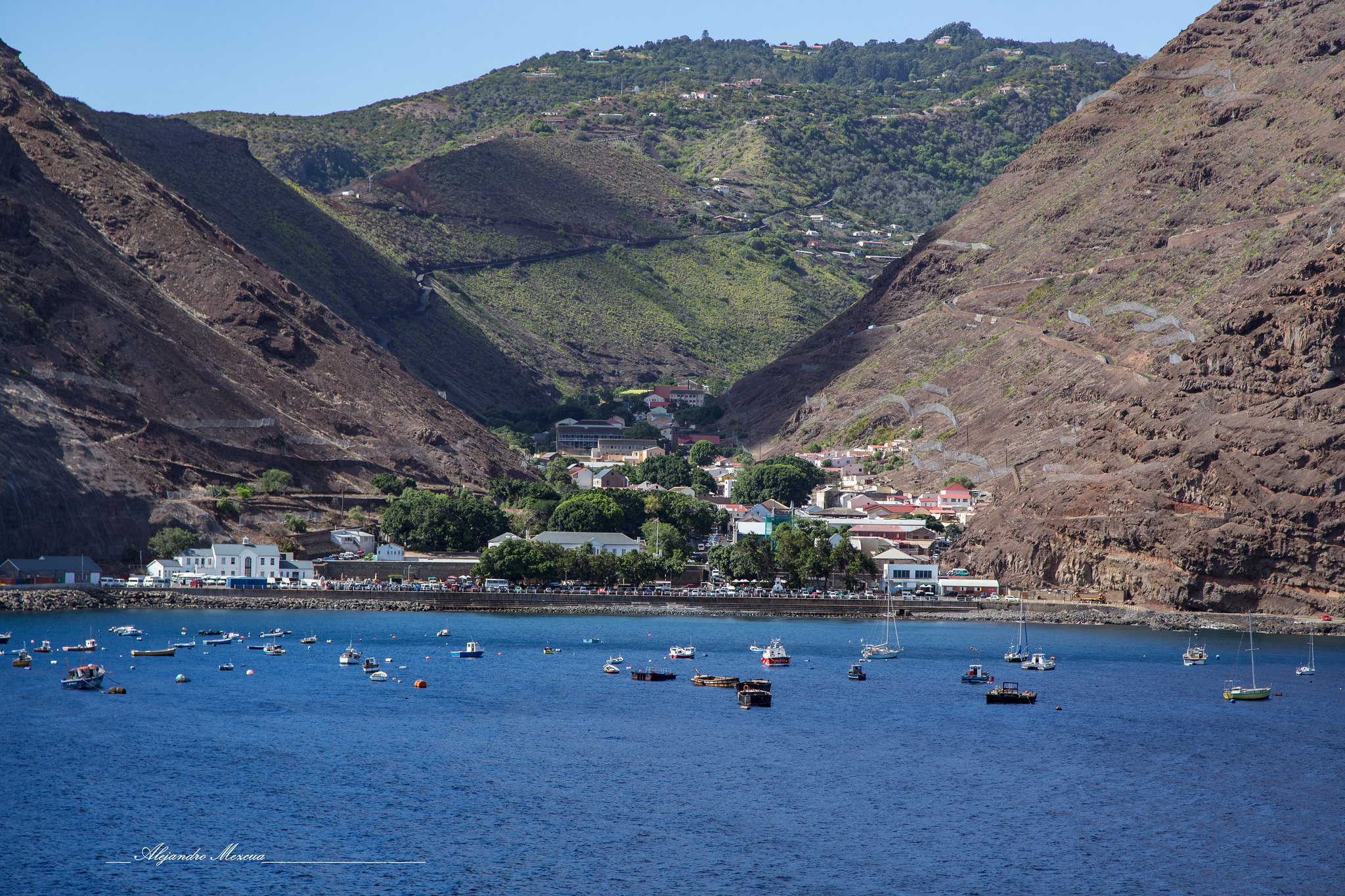 A view from the sea looking into a narrow valley on the rugged coast of St Helena. The town of Jamestown is nestled between two steep, brown, rocky cliffs, with colorful buildings and green trees concentrated in the valley floor. Small boats and yachts are anchored in the calm, deep blue waters of James Bay in the foreground.