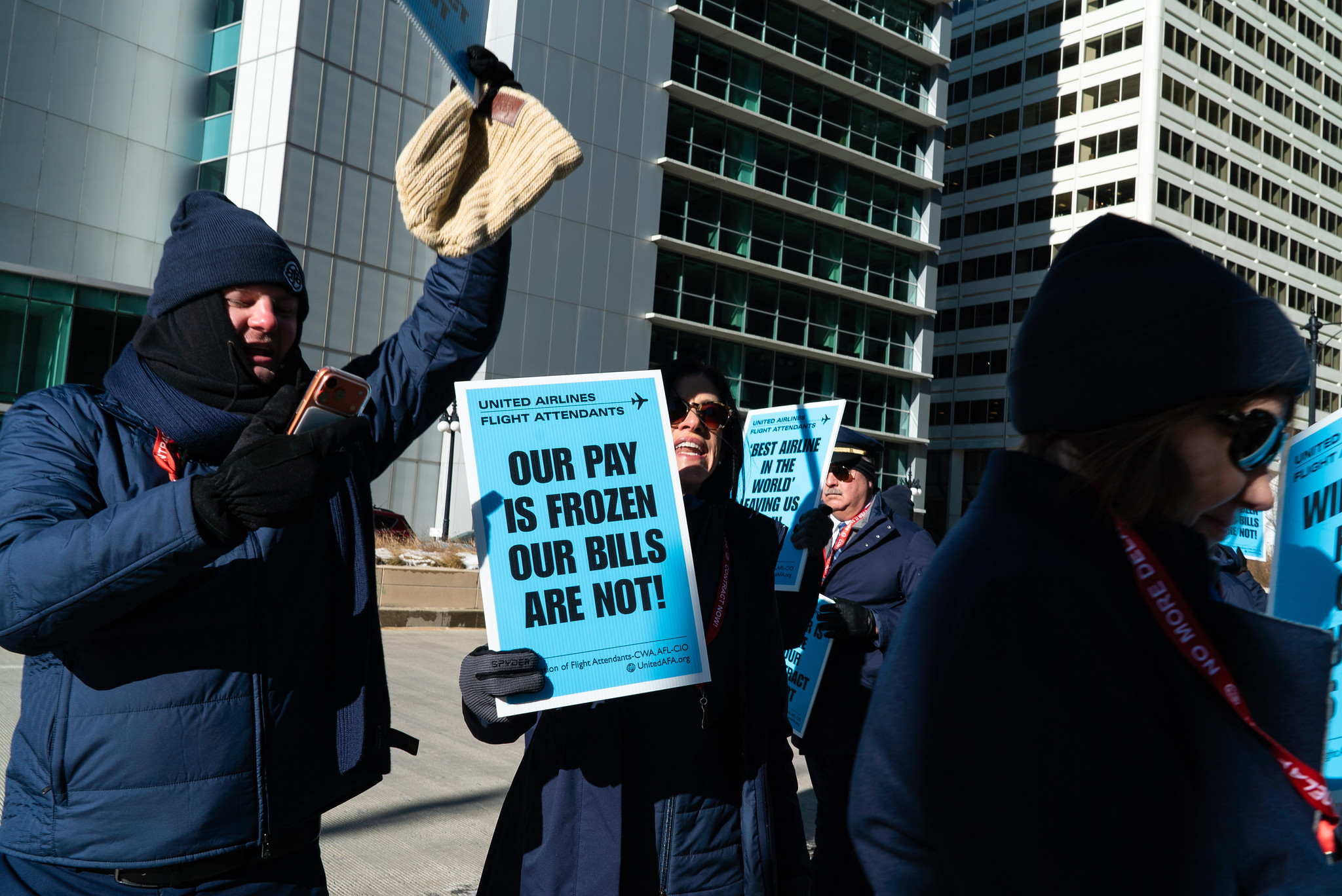 United Airlines flight attendants in winter coats and hats stand outside a tall office building during a protest. The central figure holds a bright blue sign that reads 
