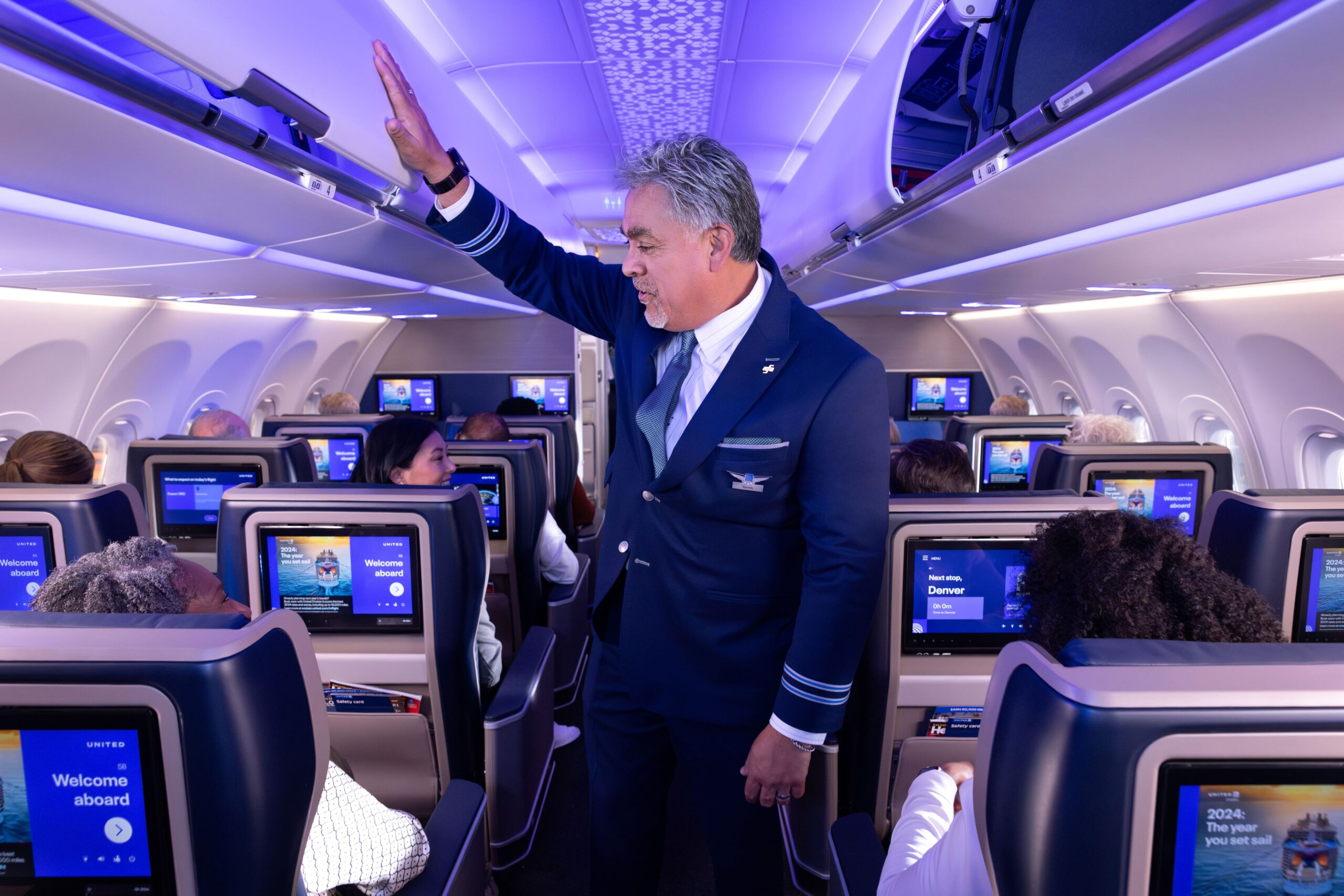 Interior view of a United Airlines cabin featuring a flight attendant in a dark blue uniform standing in the aisle, reaching up to close or assist with an overhead bin. Passengers are seated in modern blue seats equipped with large seatback entertainment screens displaying 
