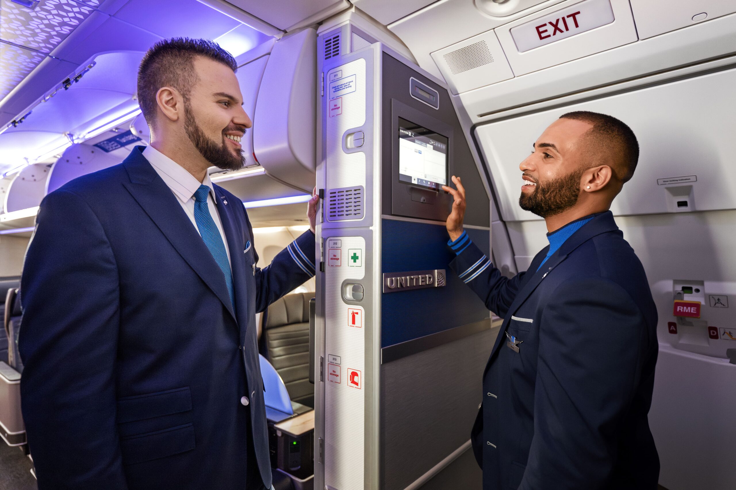 Two male United Airlines flight attendants in dark blue uniforms stand inside an aircraft cabin near the galley. One is smiling while the other interacts with a touchscreen control panel mounted on a bulkhead labeled with the United logo. The cabin is illuminated with purple ambient lighting.