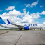A side profile of a United Airlines Boeing 737 MAX 8 parked on a concrete airport taxiway under a bright blue sky with scattered white clouds. The aircraft features the modern "Evolution" livery with a blue tail globe, blue engine nacelles, and "UNITED" in large blue lettering across the white fuselage.