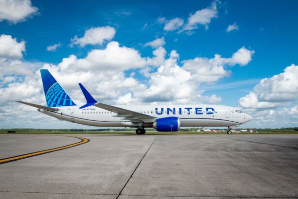 A side profile of a United Airlines Boeing 737 MAX 8 parked on a concrete airport taxiway under a bright blue sky with scattered white clouds. The aircraft features the modern "Evolution" livery with a blue tail globe, blue engine nacelles, and "UNITED" in large blue lettering across the white fuselage.