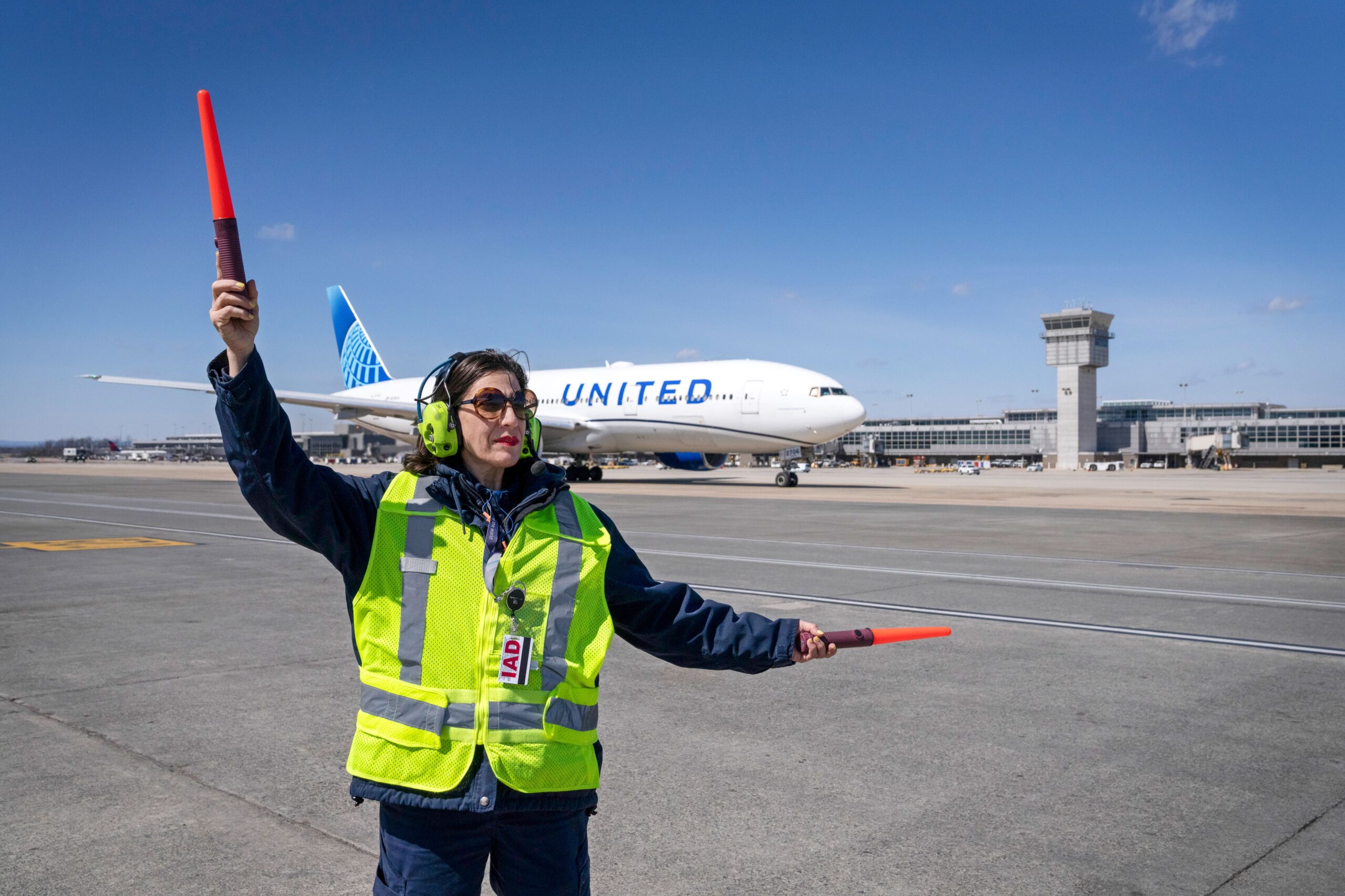 A female ramp agent wearing a high-visibility yellow vest, noise-canceling headphones, and sunglasses stands on the tarmac holding two orange marshaling wands. In the background, a large United Airlines wide-body jet sits parked near an airport terminal and a control tower under a clear blue sky.