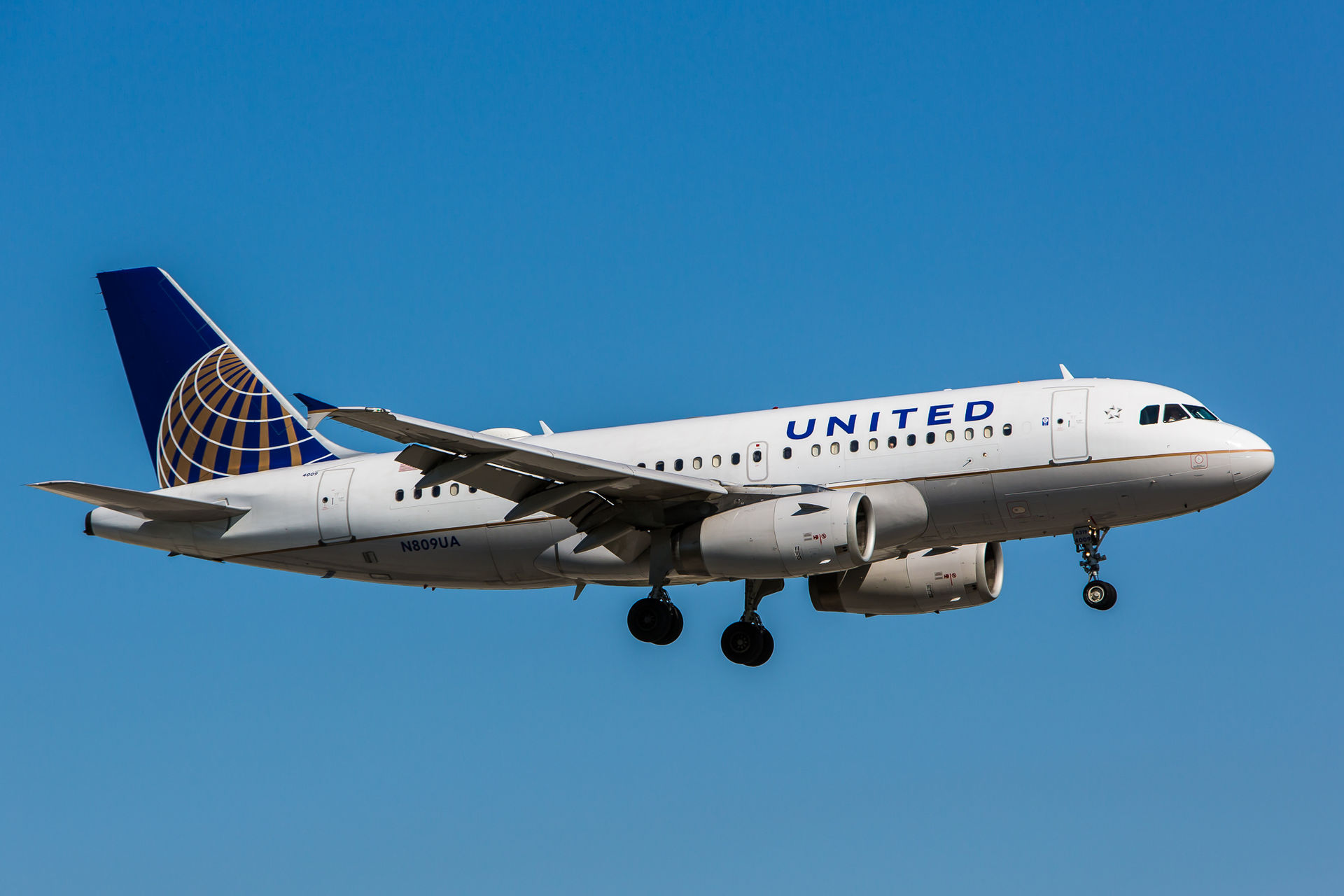 A white and blue United Airlines passenger jet is captured from a side-on, slightly lower angle as it flies through a cloudless blue sky. The aircraft features the blue 