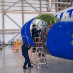 Two aircraft technicians in dark blue uniforms work on a large jet engine inside a bright, spacious hangar. One technician stands on a ladder inspecting the internal components, while the other stands on the ground supporting the open blue engine cowling. The tail end of the United Airlines plane, featuring its signature blue livery, is visible in the background.