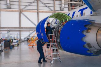 Two aircraft technicians in dark blue uniforms work on a large jet engine inside a bright, spacious hangar. One technician stands on a ladder inspecting the internal components, while the other stands on the ground supporting the open blue engine cowling. The tail end of the United Airlines plane, featuring its signature blue livery, is visible in the background.