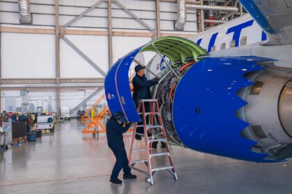 Two aircraft technicians in dark blue uniforms work on a large jet engine inside a bright, spacious hangar. One technician stands on a ladder inspecting the internal components, while the other stands on the ground supporting the open blue engine cowling. The tail end of the United Airlines plane, featuring its signature blue livery, is visible in the background.