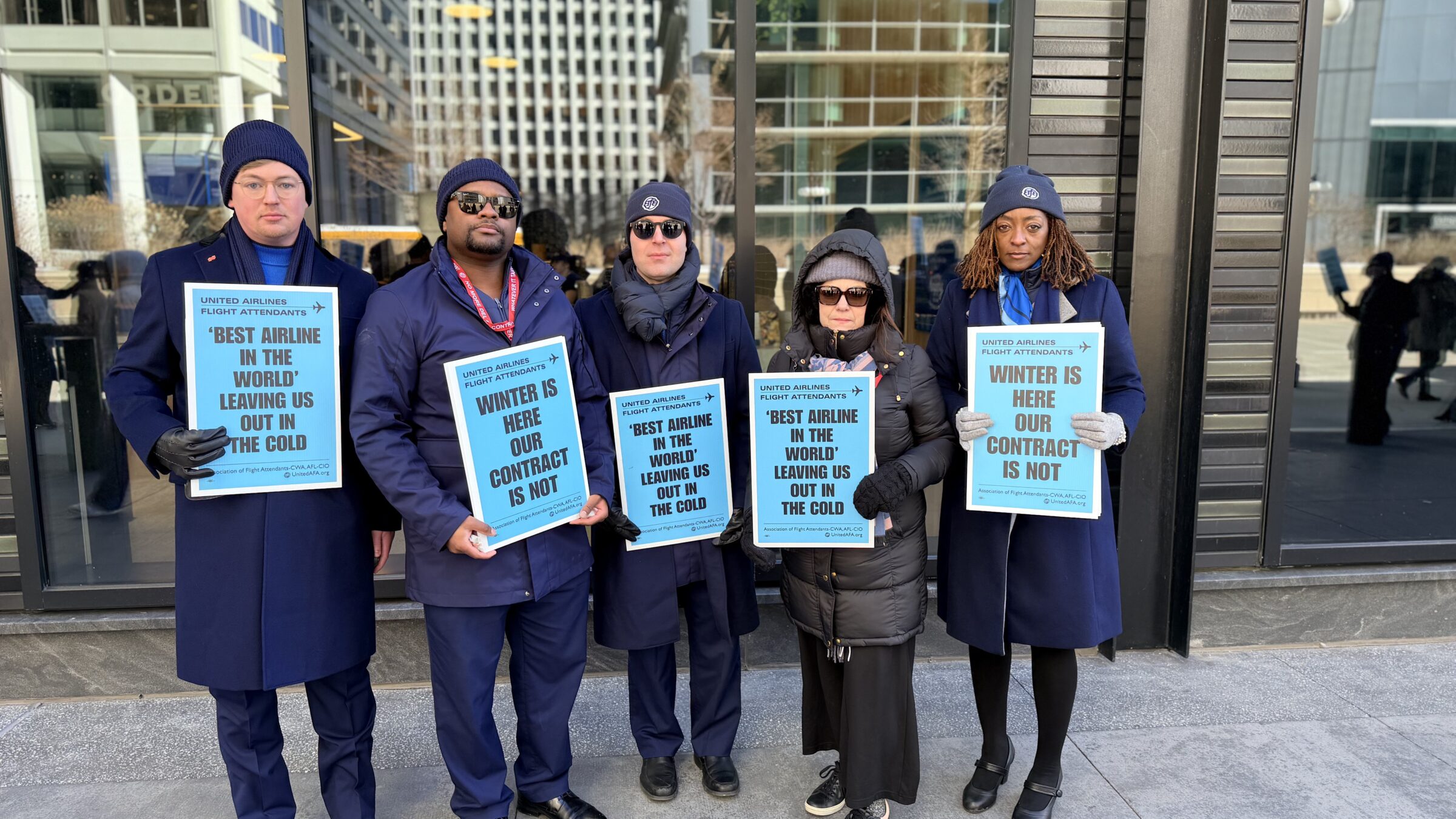 Five United Airlines flight attendants stand in a row outdoors during a winter protest. They are dressed in heavy dark coats and beanies, holding matching teal signs that read 
