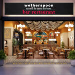 The storefront of the "Castell de Santa Bàrbera" pub, the first JD Wetherspoon location in mainland Europe, situated in the airside departures area of Alicante-Elche Miguel Hernández Airport. The entrance features a large blue sign with the pub's name in white lettering, located next to an airport restroom and opposite gate B30.
