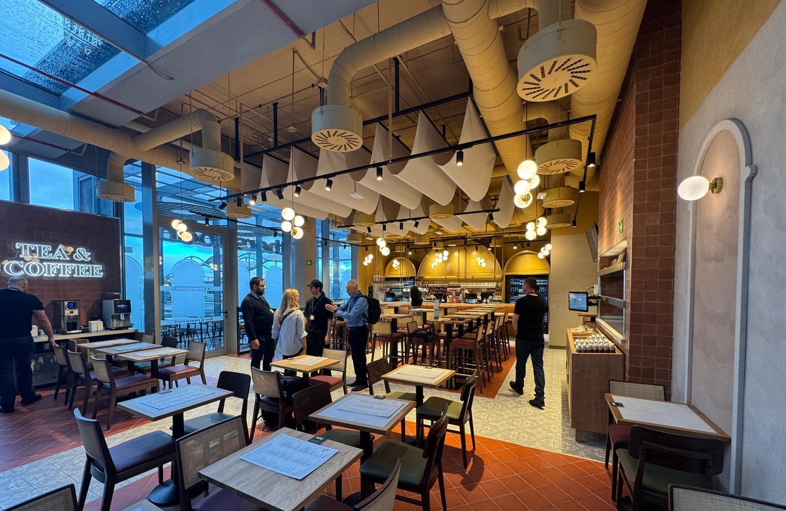 The interior of a modern airport restaurant with high ceilings and industrial-style exposed ducting. Large windows on the left look out onto the airfield. The space is furnished with wooden tables and chairs, a 