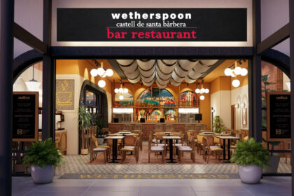 The storefront of the "Castell de Santa Bàrbera" pub, the first JD Wetherspoon location in mainland Europe, situated in the airside departures area of Alicante-Elche Miguel Hernández Airport. The entrance features a large blue sign with the pub's name in white lettering, located next to an airport restroom and opposite gate B30.