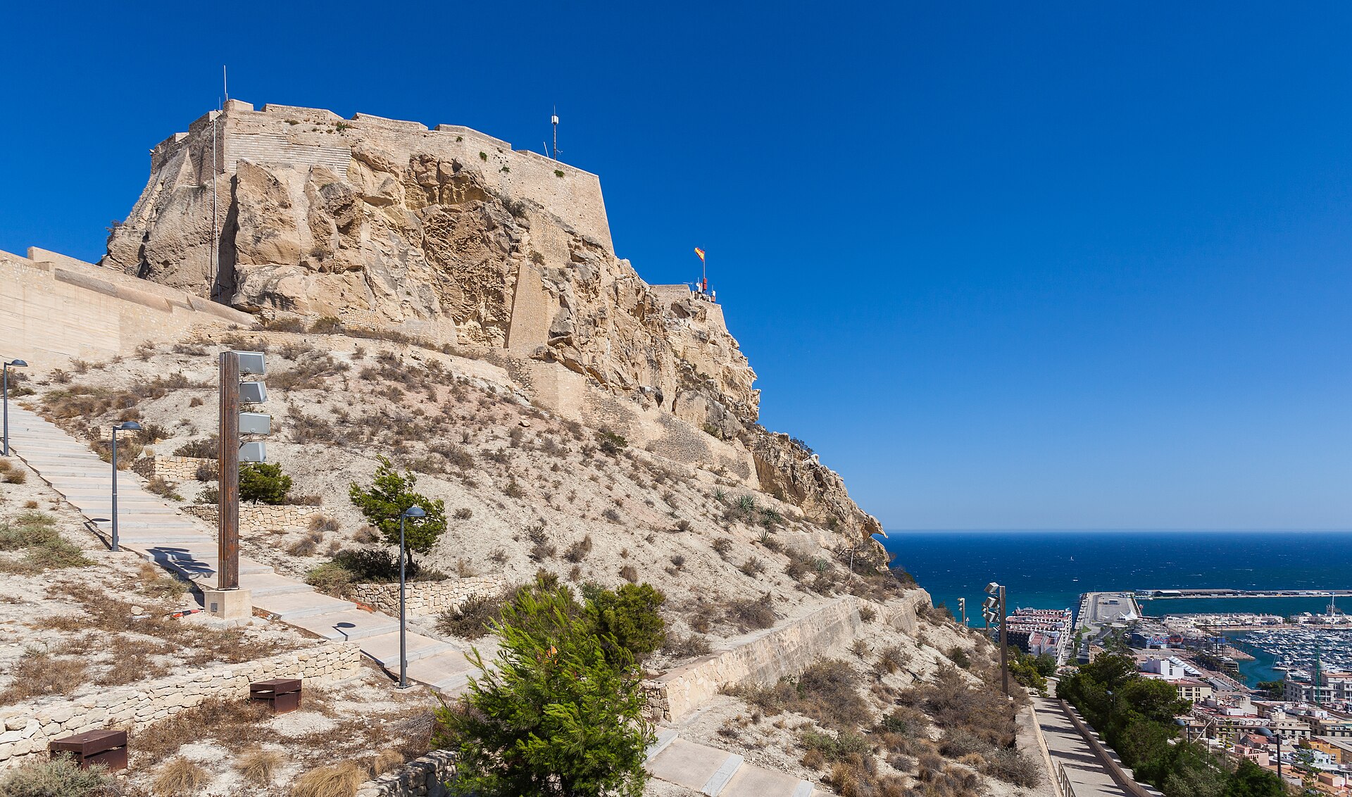 A view of the historic Castell de Santa Bàrbera, a large stone fortification atop a rocky mountain in Alicante, Spain. A paved stone pathway with modern lampposts winds up the arid hillside toward the fortress. In the background, the deep blue Mediterranean Sea and the Alicante harbor filled with boats are visible under a clear, bright sky.