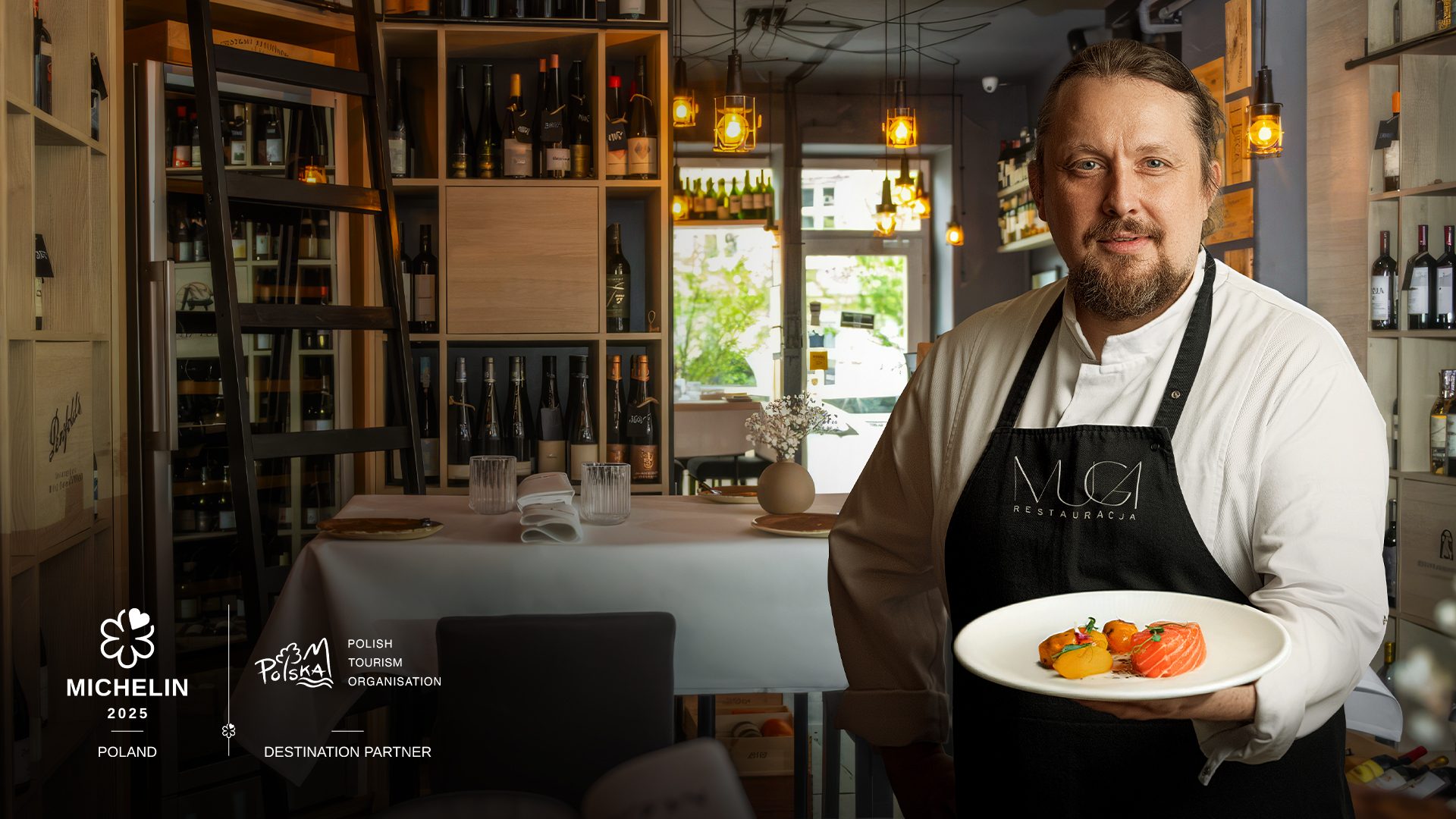 A Michelin-starred chef holding a plated dish inside a restaurant dining room, photographed at Muga restaurant in Poznań.