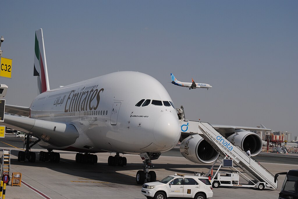 Emirates Airbus A380-800 parked at a gate labelled C32 at Dubai International Airport. Sky above, a smaller aeroplane captured mid-flight.