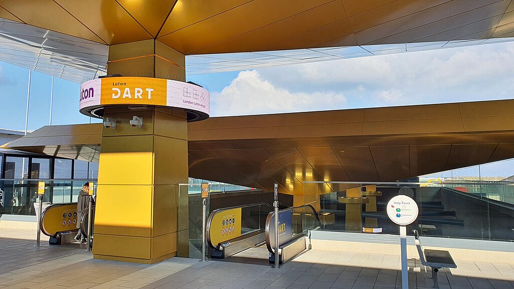 Entrance to the Luton DART station at London Luton Airport, with golden architectural panels. Two escalators lead downward to the train platforms, and a help point kiosk stands to the right