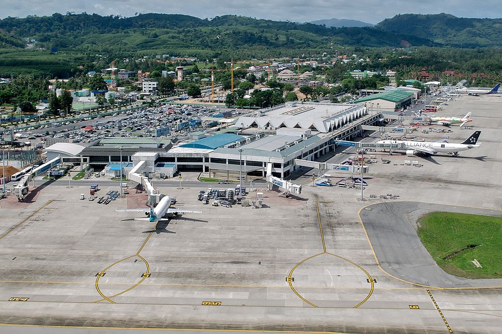 Aerial view of Phuket Airport; several aircraft on the tarmac; green hills in the background.