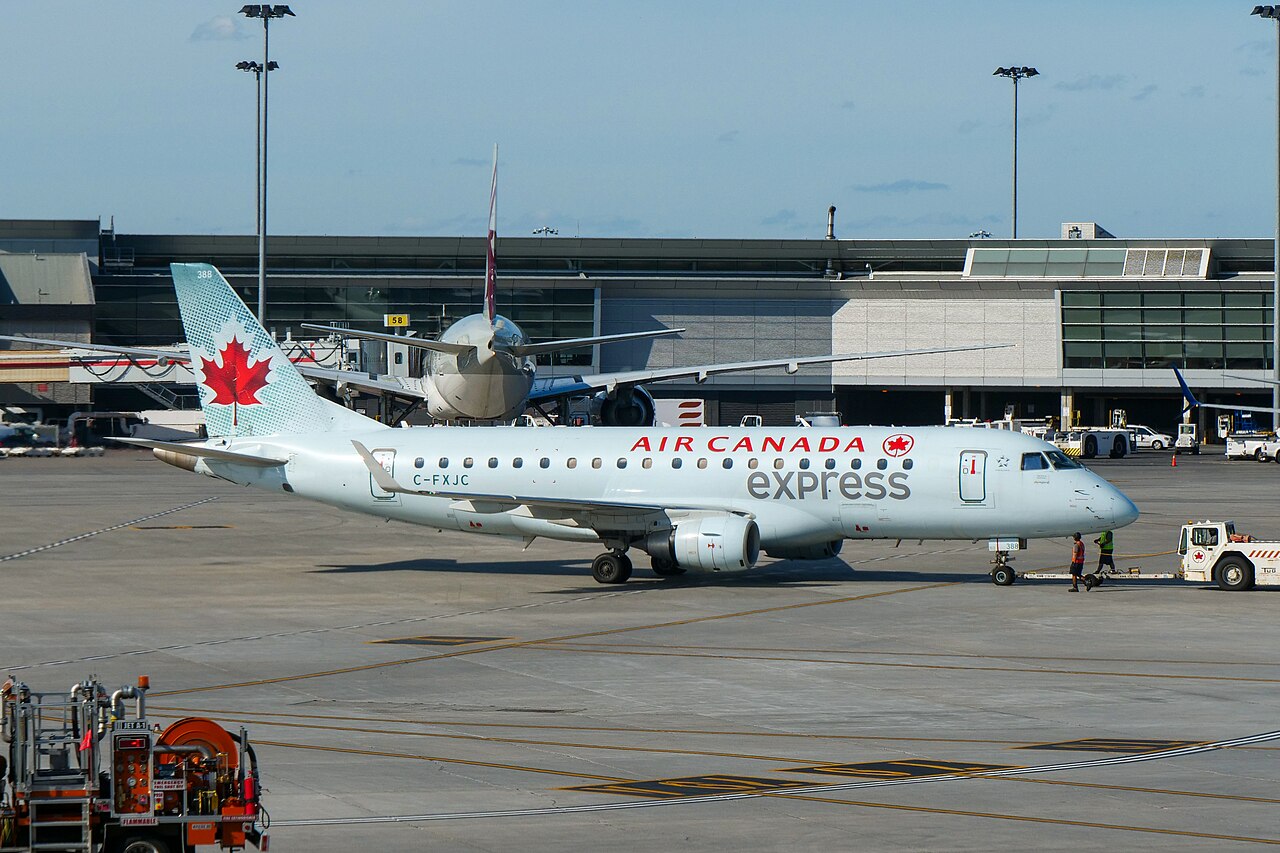 A white regional passenger jet with “Air Canada Express” branding sits on an airport apron, being pushed back by a small ground tug.