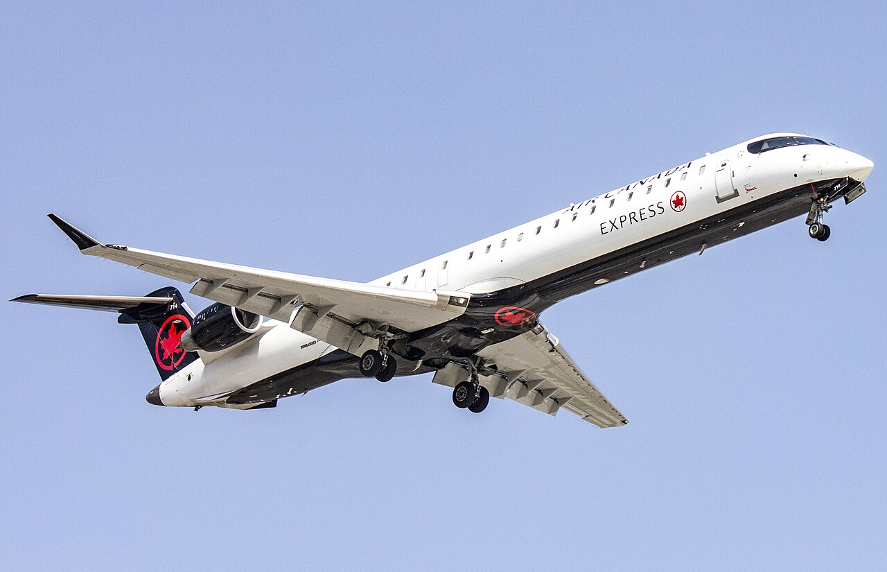 A white regional passenger jet in mid-flight climbs against a clear blue sky, captured from below at an angle that shows the underside of its wings and fuselage.