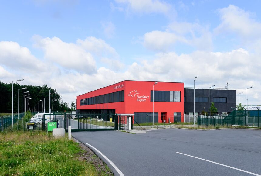 A bright Red building named " Frankfurt Airport" stands behind a fenced entrance.