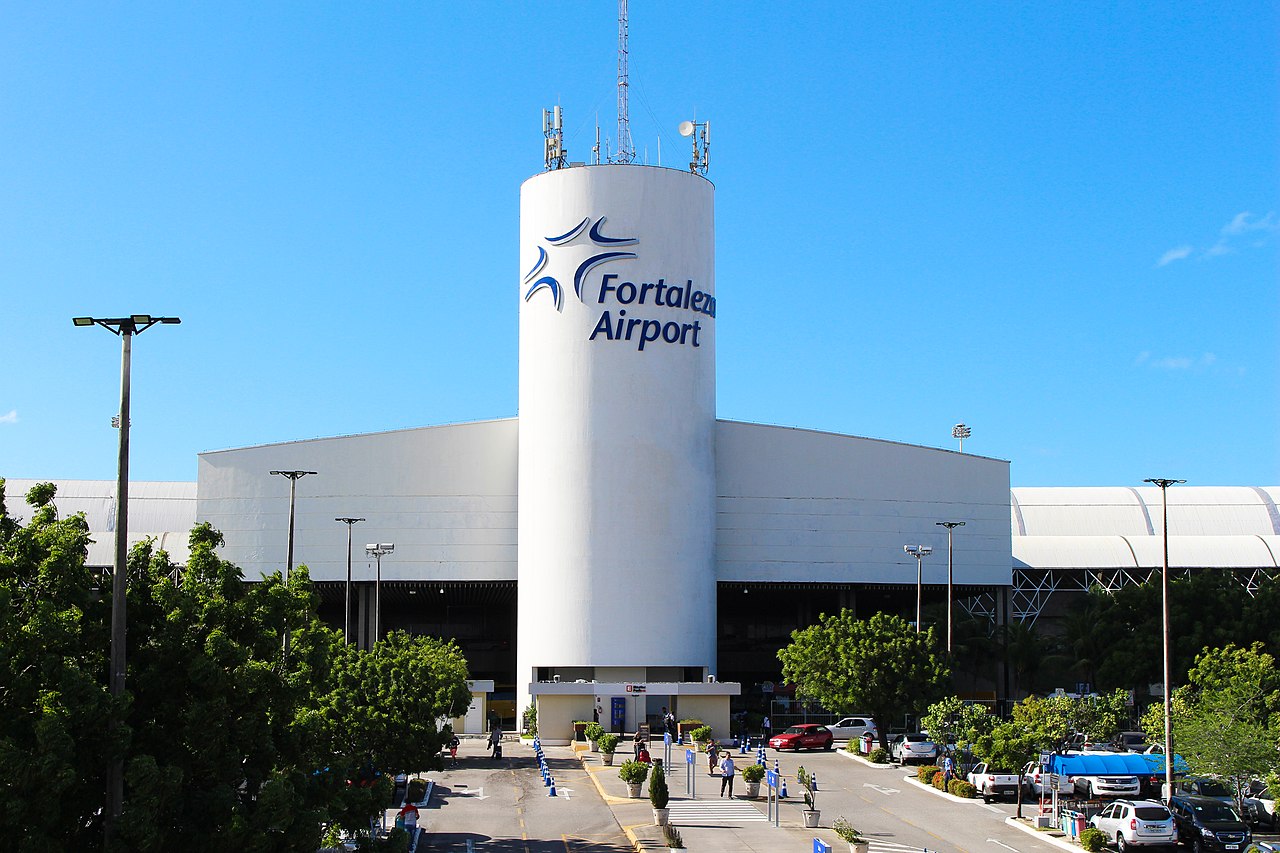 A modern white terminal building at Fortaleza Airport stands under a clear blue sky, featuring a tall cylindrical structure with the airport’s name and logo displayed near the top.